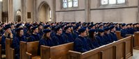Graduation ceremony in gothic cathedral with students in blue caps and gowns seated in wooden pews during commencement celebration.