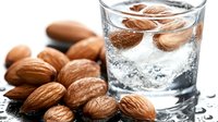 Fresh almonds soaking in pure water with ice cubes in glass, raw nuts scattered on dark surface with water drops, macro food photography.