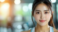 Young Asian woman with natural makeup and gentle smile against blurred bokeh background, wearing casual blue tank top, radiating confidence and warmth.