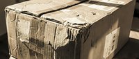 Damaged cardboard shipping box with torn edges and worn surface showing wear from handling and transport in warehouse or delivery setting.