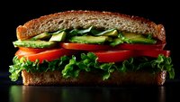 Fresh vegetable sandwich with layers of crisp lettuce, ripe tomatoes, and sliced avocado between whole grain bread slices on dark background with dramatic lighting.