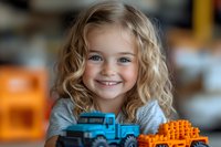 Young blonde girl with blue eyes and curly hair smiling brightly while playing with colorful toy truck and building blocks, natural indoor lighting creates warm atmosphere.