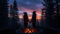 Two people holding hands by campfire at sunset in forest with dramatic sky and pine trees surrounding them.