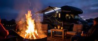 RV camping scene with crackling campfire at twilight, motorhome parked nearby under dramatic purple sky. Outdoor adventure and recreational vehicle lifestyle.