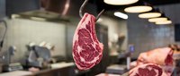 Raw beef steak hanging in professional butcher shop with blurred kitchen background, showcasing quality meat selection for culinary use.