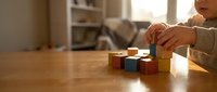 Child playing with colorful wooden building blocks on table in cozy home interior, developing fine motor skills and creativity through educational toy play.