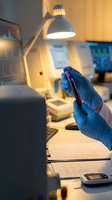 Lab technician in blue gloves holds blood sample tube in clinical laboratory with medical equipment and monitors in background.