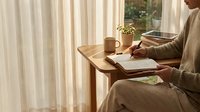 Woman in cozy beige sweater writing in journal at wooden desk by window with warm morning light, coffee cup and plant creating peaceful workspace atmosphere.