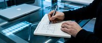Architect or engineer in dark business suit sketching building blueprint on paper at glass desk with laptop and modern office environment.