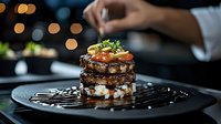 Gourmet layered dish with grilled meat and fresh garnish being prepared by chef in upscale restaurant with bokeh lights in background.
