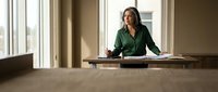 Professional businesswoman in green blazer sitting at conference table in modern office boardroom with large windows and natural lighting.
