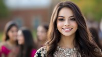 Young Hispanic woman with radiant smile wearing embellished formal dress at outdoor celebration, with blurred people in background. Natural daylight portrait.