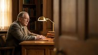 Senior man with glasses working at wooden desk with brass lamp in traditional library office setting with warm lighting and bookshelves.