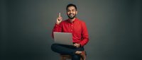 Young Middle Eastern businessman in red shirt pointing up with laptop, sitting on stool against dark background. Professional male entrepreneur with idea gesture.
