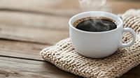 Steaming hot black coffee in white ceramic cup placed on knitted beige coaster against rustic wooden table background, creating cozy morning atmosphere.