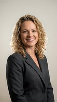Professional businesswoman with curly blonde hair smiling confidently in dark suit jacket against neutral background for corporate headshot.