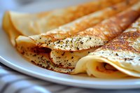 Thin golden crepes sprinkled with black poppy seeds and spices on white plate, close-up view showing delicate texture and crispy edges of traditional breakfast pancakes.