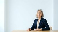 Mature woman with white hair and light skin tone, wearing dark navy blazer and white blouse, seated at wooden desk against white background for corporate and business use.