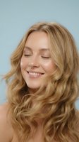 Young blonde woman with wavy hair smiling with eyes closed showing natural beauty and joy against soft blue background for wellness and lifestyle content.