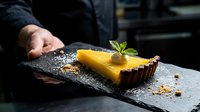 Professional chef presenting elegant lemon tart dessert on dark slate plate with mint garnish and crumb details in upscale restaurant kitchen setting.