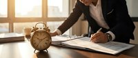 Professional businessman in dark suit reviewing documents at modern office desk with vintage alarm clock, emphasizing time management and productivity concepts.