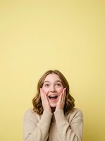 Excited young woman with shoulder-length brown wavy hair and fair complexion wearing beige sweater, hands on cheeks, expressing surprise and joy against yellow background for advertising and lifestyle content.
