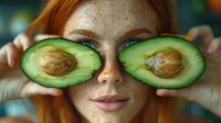 Young redhead woman with freckles holding fresh cut avocado halves over eyes during beauty skincare routine, natural organic facial treatment concept.