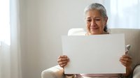 Senior woman with gray hair and South Asian ethnicity holding blank white poster while sitting on couch indoors for advertising mockup.