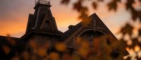 Victorian Gothic mansion with ornate architecture and bell tower silhouetted against dramatic orange sunset sky through autumn foliage frame.