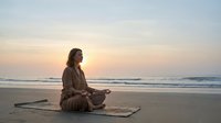 Young Caucasian woman meditating in lotus pose on beach at sunrise, practicing mindfulness and yoga by the ocean for wellness.