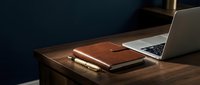 Leather notebook and pen on wooden desk beside laptop in dark blue office, professional workspace setup for business and productivity.