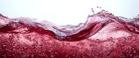 Dynamic red liquid wave with splashing droplets and bubbles against light background, creating dramatic motion and texture.