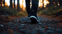 Person walking on rocky forest path during golden sunset, close-up view of black sneaker and jeans. Atmospheric autumn outdoor scene with bokeh light effects.