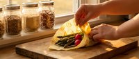 Fresh vegetables wrapped in yellow cloth on wooden cutting board, hands holding bundle, glass storage jars with grains in background, natural kitchen lighting, sustainable food storage and meal prep.