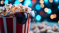 Fresh popcorn overflowing from classic red striped container with sunglasses against blurred bokeh lights background, creating cinematic atmosphere.