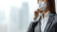 Asian businesswoman wearing protective face mask looking through window in office building, contemplative pose during pandemic workday.
