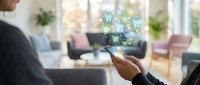 Person using smartphone for online shopping with digital cart icons floating above device in modern living room interior.