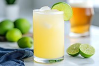 Fresh lime margarita cocktail with ice and lime wheel garnish on kitchen counter with cut limes and whiskey glass in background for summer entertaining.
