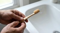 Elderly hands holding eco-friendly bamboo toothbrush over white bathroom sink promoting sustainable oral hygiene and zero waste lifestyle choices.