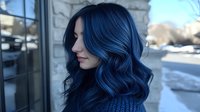Young Asian woman with vibrant blue wavy hair styled in loose curls, wearing dark knit sweater, looking away pensively against stone wall background.