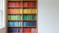 Colorful rainbow bookshelf with organized books arranged by color spectrum from red to purple in wooden library shelving unit against white brick wall.