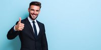 Confident businessman in dark suit giving thumbs up gesture against blue background, expressing success and positive attitude for corporate presentations.