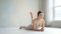 Young Caucasian woman in gray sports bra practicing yoga pose on floor in bright studio with natural light from window during morning workout session.