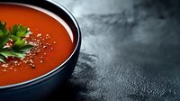 Homemade tomato soup garnished with fresh parsley and seasonings served in dark ceramic bowl against textured black background with copy space.