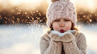 Young girl in knitted hat and mittens blowing snow from her hands against golden bokeh winter background.