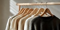 Neutral earth tone sweaters hanging on wooden hangers in minimalist closet with natural light creating shadow patterns.