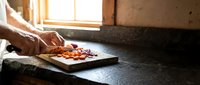 Male chef hands slicing fresh tomatoes on wooden cutting board in rustic kitchen with natural sunlight streaming through window for cooking preparation.