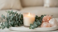 Spa wellness arrangement with burning candle, eucalyptus branches and himalayan salt stones on white tray creating peaceful home relaxation atmosphere.