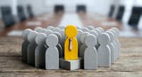 Yellow wooden figure stands out among gray figures in business leadership concept on conference table with blurred boardroom background.