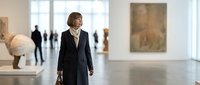 Woman in professional black blazer walking through modern art gallery or museum exhibition space with contemporary artwork and sculptures displayed.